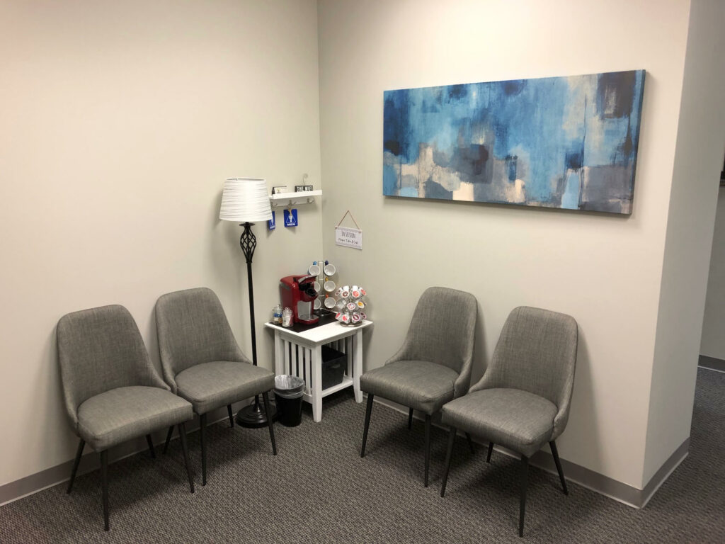 A small waiting area with four gray chairs, a coffee station on a side table, a floor lamp, and a blue abstract painting on the wall.