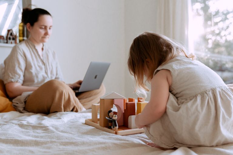 Side view of adorable toddler girl sitting on bed and playing with wooden toys while mother working from home with laptop on knees on sunny morning with working mom guilt